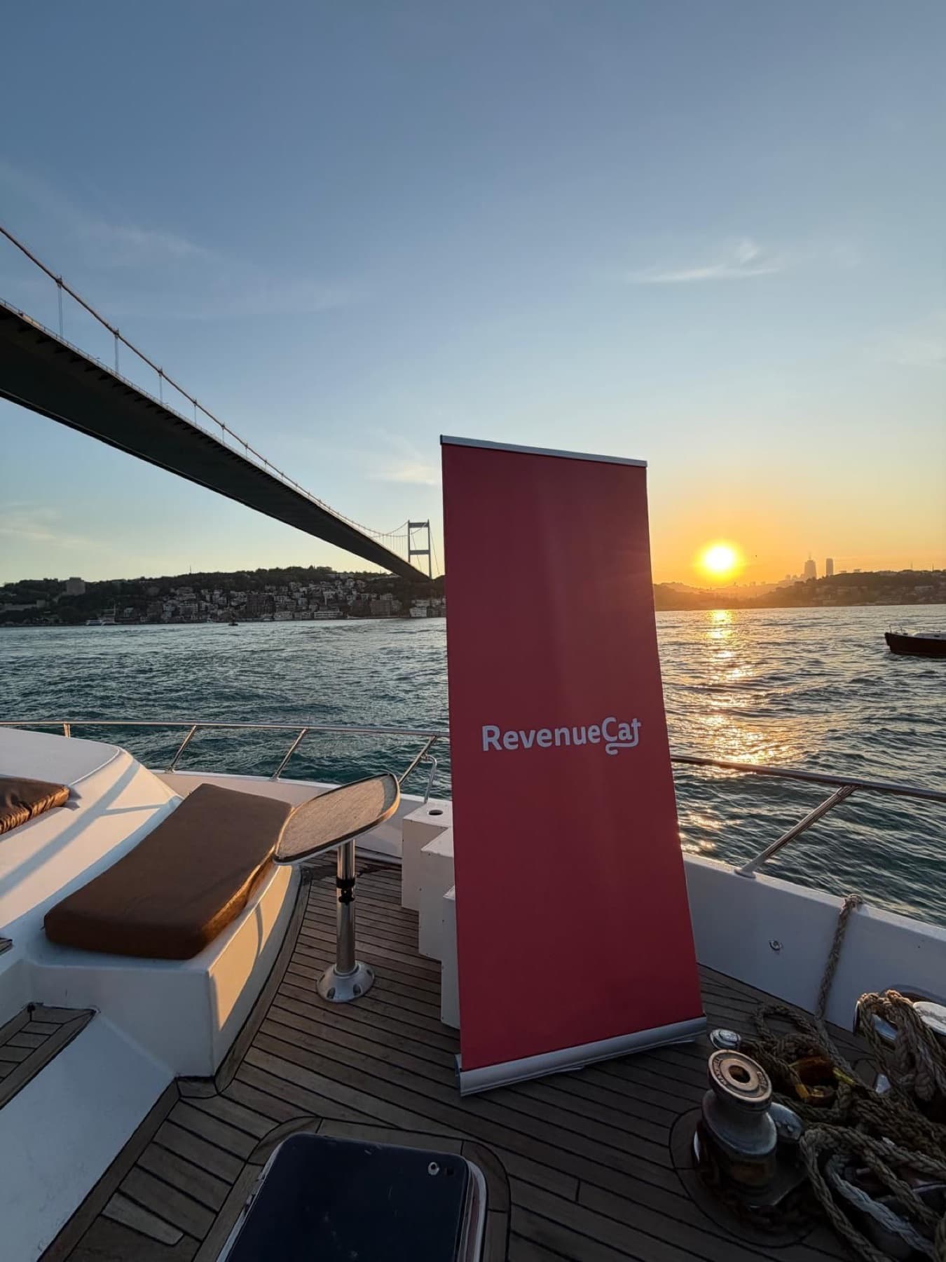 A RevenueCat banner on the deck of the App Founders Sunset Yacht Party on the Bosphorus in Istanbul, with the bridge and the sunset behind it